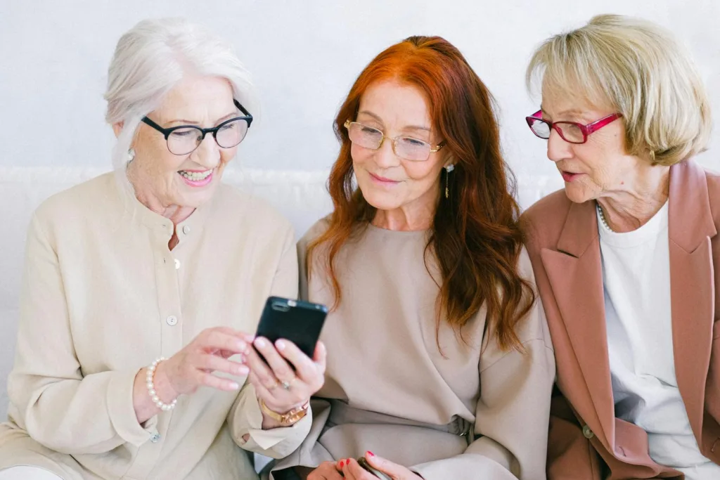 Three senior women enjoying a smartphone, sharing smiles and joy indoors.